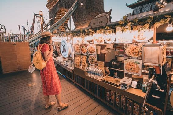 Dubai Global Village-t látogató piros pöttyös ruhába lévő nő nemzeti ételeket nézeget.