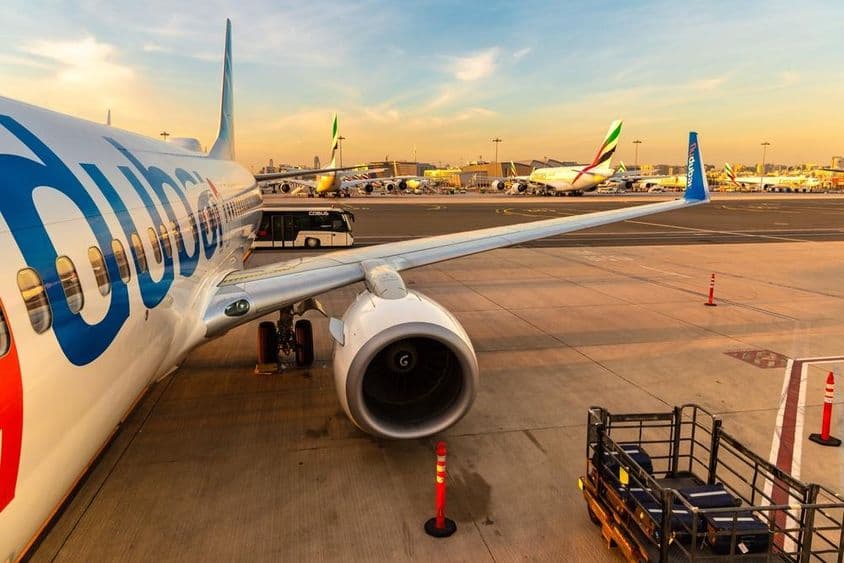 Flydubai repülő áll a parkolóhelyen, mögötte több másik repülő a háttérbe, és egy utasokat szállító busz, jobbra a csomagok egy kis szállító járműben.