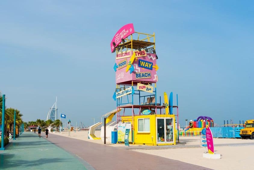 Dubai Kite Beach üdvözlőépítmény, táblákkal, háttérben a Burj Al Arab, előtte futópálya és homokos part.
