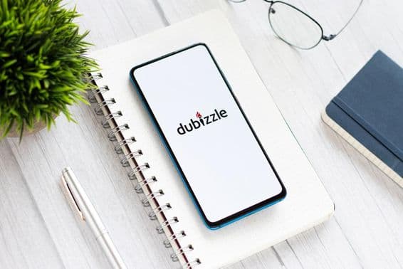 Egy asztalon egy telefon amin a dubizzle fut, alatta egy jegyzettömb, mellette egy tól szemüveg, kis zöld növény.