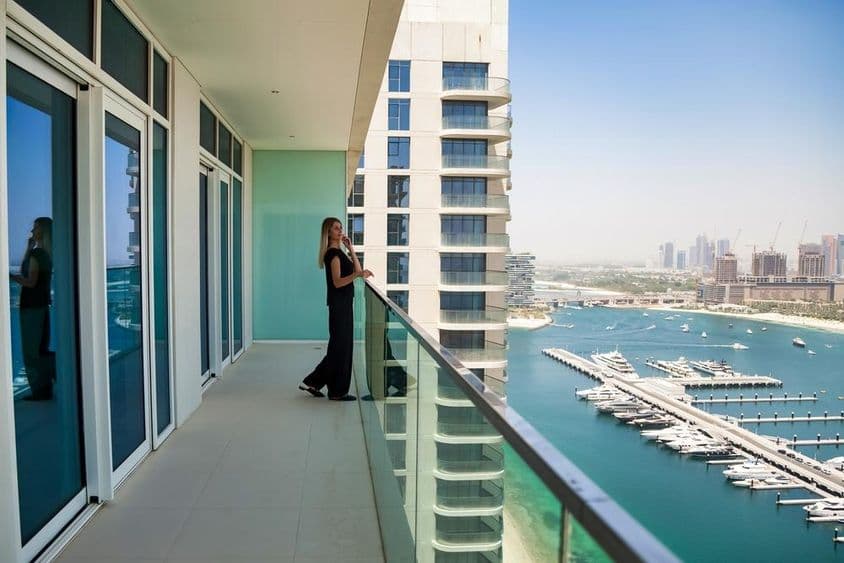Egy nőt látunk Dubai teraszon ahogy a csatornán úszkáló hajókat nézi, mögötte egy felhőkarcoló látható.