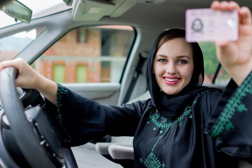Egy Dubai hölgy kezében egy jogosítványt tart, másik kezével a kormányt fogja, mosolyog.