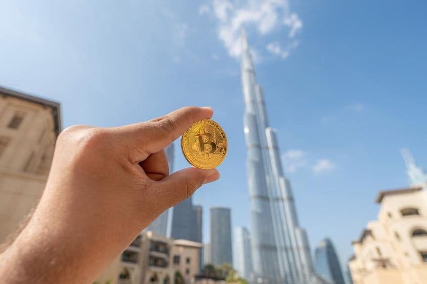 Arany bitcoin érmét tart a kezében egy ember, égnek fordítja , háttérben a burj khalifa.