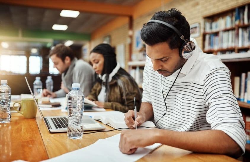 Egyetemista diákok írnak papíron, előttük könyv és notebook, egyik fején egy fülhallgató .