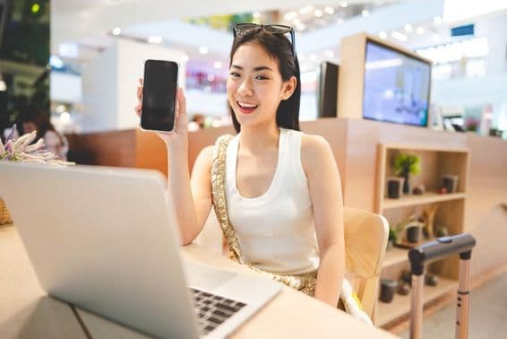 Gen Z lány dolgozik, kezében telefon, előtte notebook, mellette egy gurulós bőrönd fogantyú.