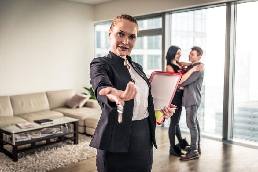 Egy női ingatlanbróker kezével hívogat , mögötte egy pár ölelkezik.