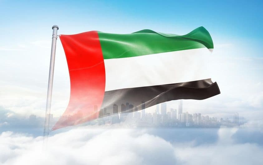 Egyesült Arab Emírségek zászlaja hullámzik a felhőkben, felhőkarcolok a háttérben.
