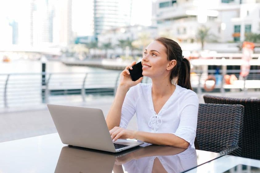 Fiatal szabadúszó nő dolgozik laptopon és ül a kávézóban a szabadban, kezében telefon.