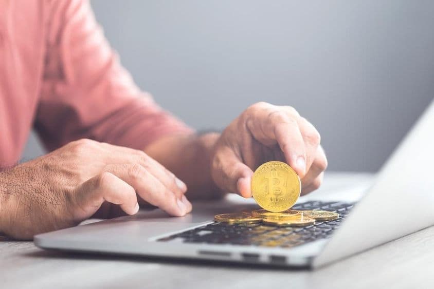 A férfi egy aranyszínű bitcoint tart egy laptop használata közben.