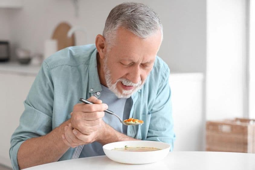 Parkinson-szindrómában szenvedő idős férfi a konyhában az asztalnál eszik.