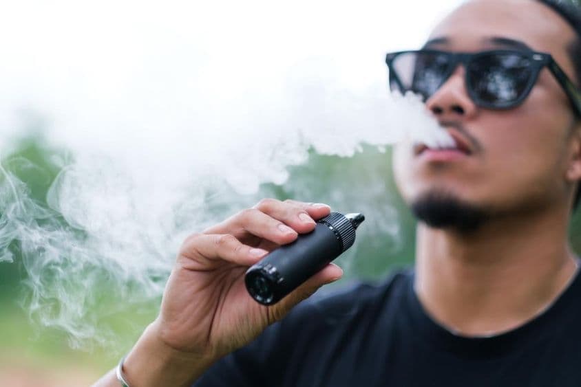 Ázsiai férfi vaping e-cigarettával dohányzik, fújja a füstöt a szájából, és élvezi a szabadban nyáron.