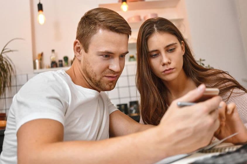 Fiatal pár, amint kávézik a konyhában, mobiltelefont használva online banki értesítést olvas.