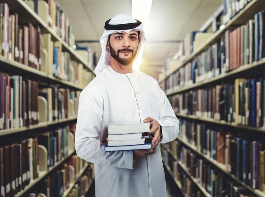 Arab diák áll a könyvtárban kezében könyvekkel.