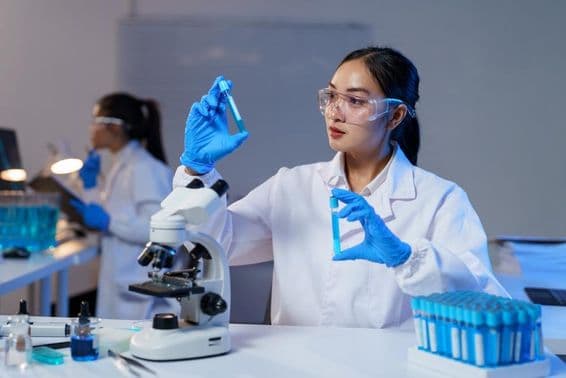 Ázsiai női tudós laborköpenyben és kesztyűben két kémcsövet hasonlít össze.