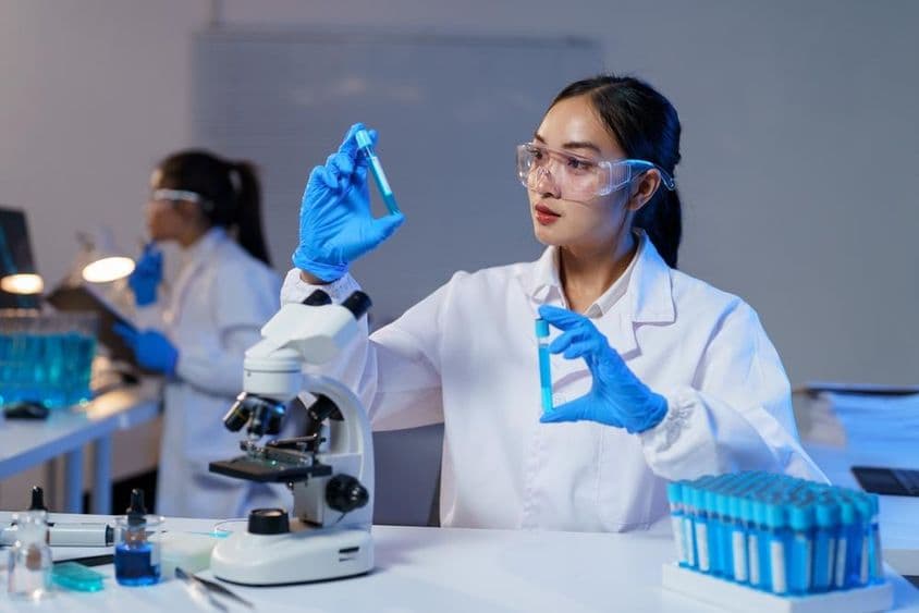 Ázsiai női tudós laborköpenyben és kesztyűben két kémcsövet hasonlít össze.