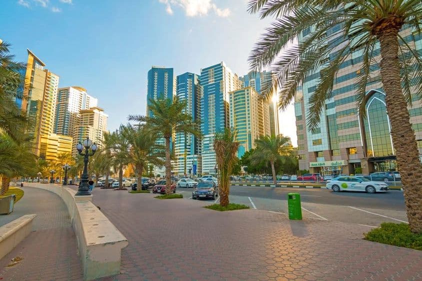 Sharjah város látképe napkeltekor, a modern felhőkarcolókra visszaverődő arany fénnyel.