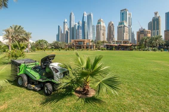 Fűnyíró traktor egy zöld mezőn a Dubai Marina felhőkarcolói előtt.