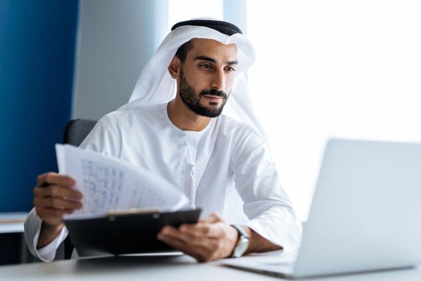 Férfi laptoppal dolgozik dubai irodájában, hagyományos fehér emirátusi ruhában.