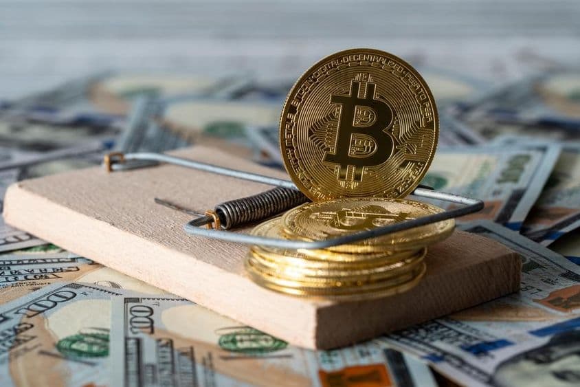 Arany kriptovaluta érmék bitcoin egérfogóban, amerikai 100 dolláros bankjegyek hátterében.