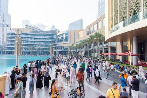 Turisták és helyiek sokszínű tömege sétál a Dubai Mall előtti forgalmas vízparti sétányon.